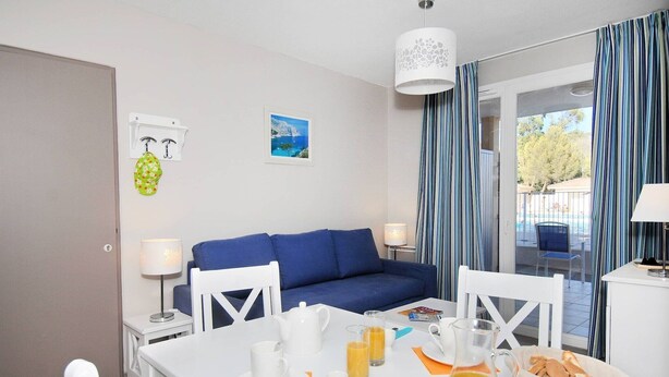 A hotel room with a blue sofa, a dining table set with breakfast items, and a view of a pool through the curtains. - Apartment in France with Pool & Pine Views (La Londe les Maures)