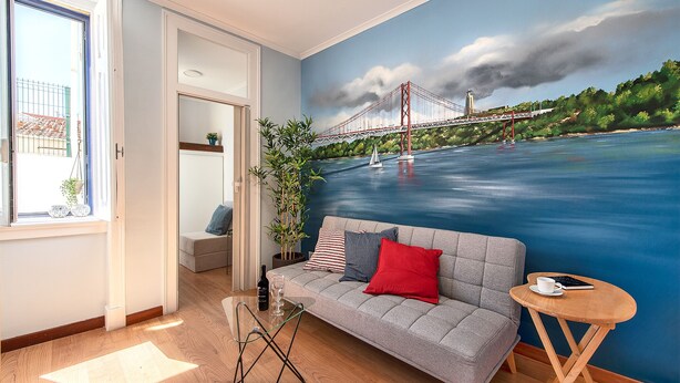 Un salon avec une grande fresque représentant un pont, un canapé avec des coussins, une petite table sur laquelle se trouvent une tasse et un livre, et une plante.