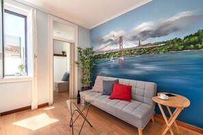 Un salon avec une grande fresque représentant un pont, un canapé avec des coussins, une petite table sur laquelle se trouvent une tasse et un livre, et une plante.