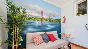 Une pièce décorée d’une grande fresque représentant un pont, avec un canapé orné de coussins à motifs, une plante en pot et une table en verre sur laquelle se trouvent des verres à vin et une bouteille.