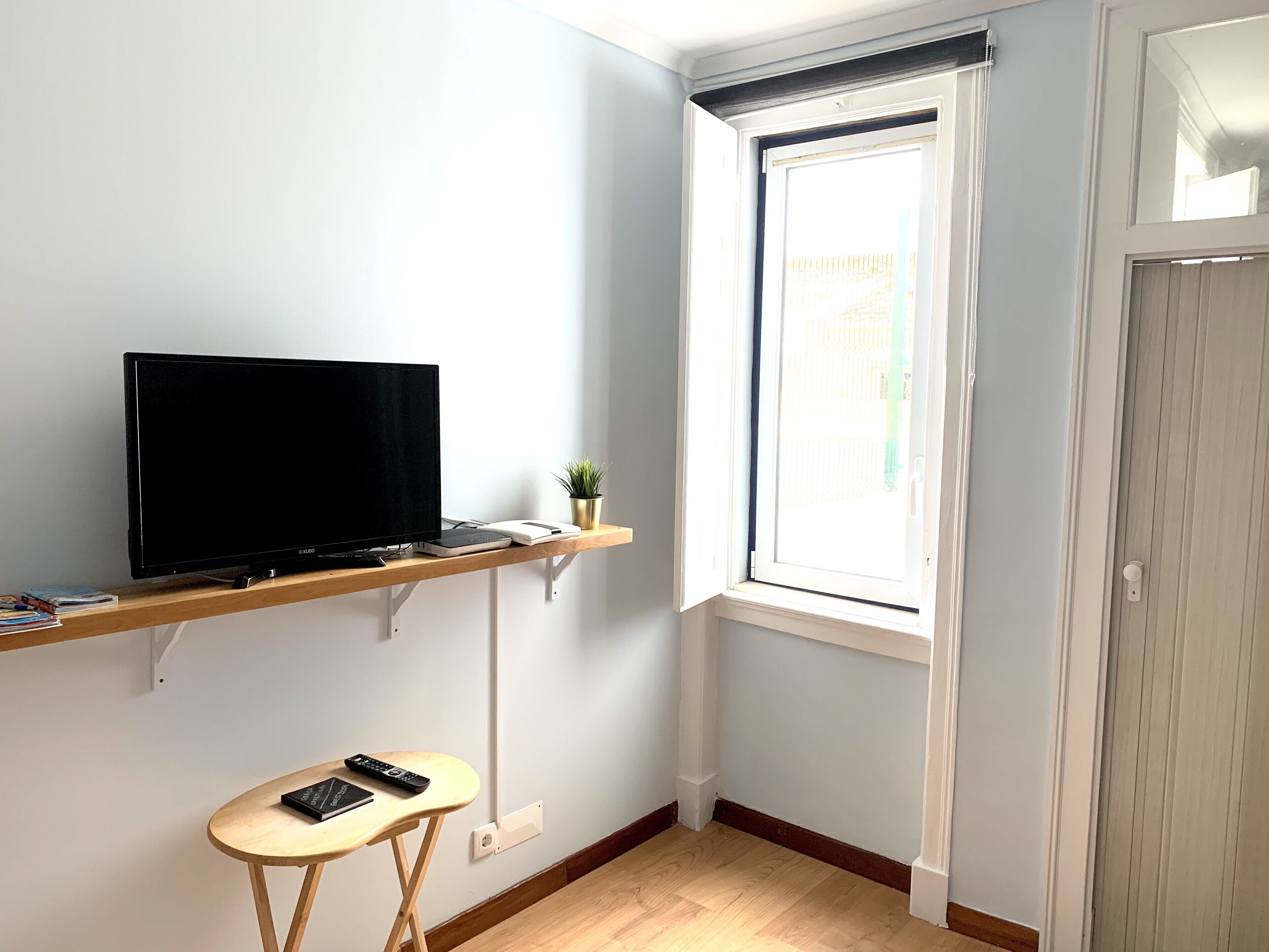 Una habitación pequeña con una televisión sobre una estantería de madera, una mesita con un libro y un mando a distancia, y una ventana con persianas.