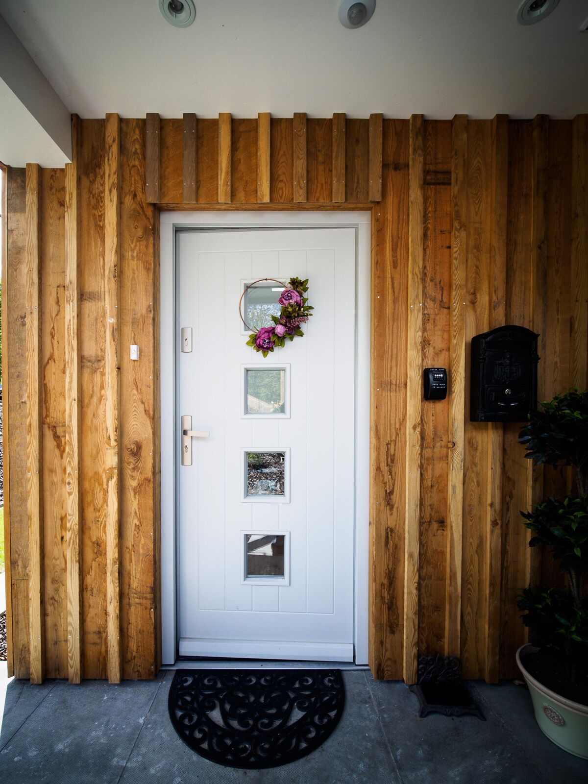 Una puerta blanca con un adorno floral, tres cuadros enmarcados y un buzón negro sobre una pared de madera.