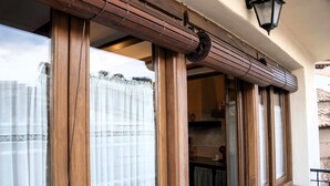Ein rahmenförmiges Holzfenster mit Vorhang, im Hintergrund eine Küche erkennbar, und ein Balkon mit Geländer.