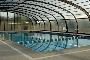 An indoor swimming pool with a curved glass ceiling, surrounded by a tiled floor.