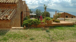A rustic building with a tiled roof, a small garden with pink flowers, and a view of a distant mountain and sea.