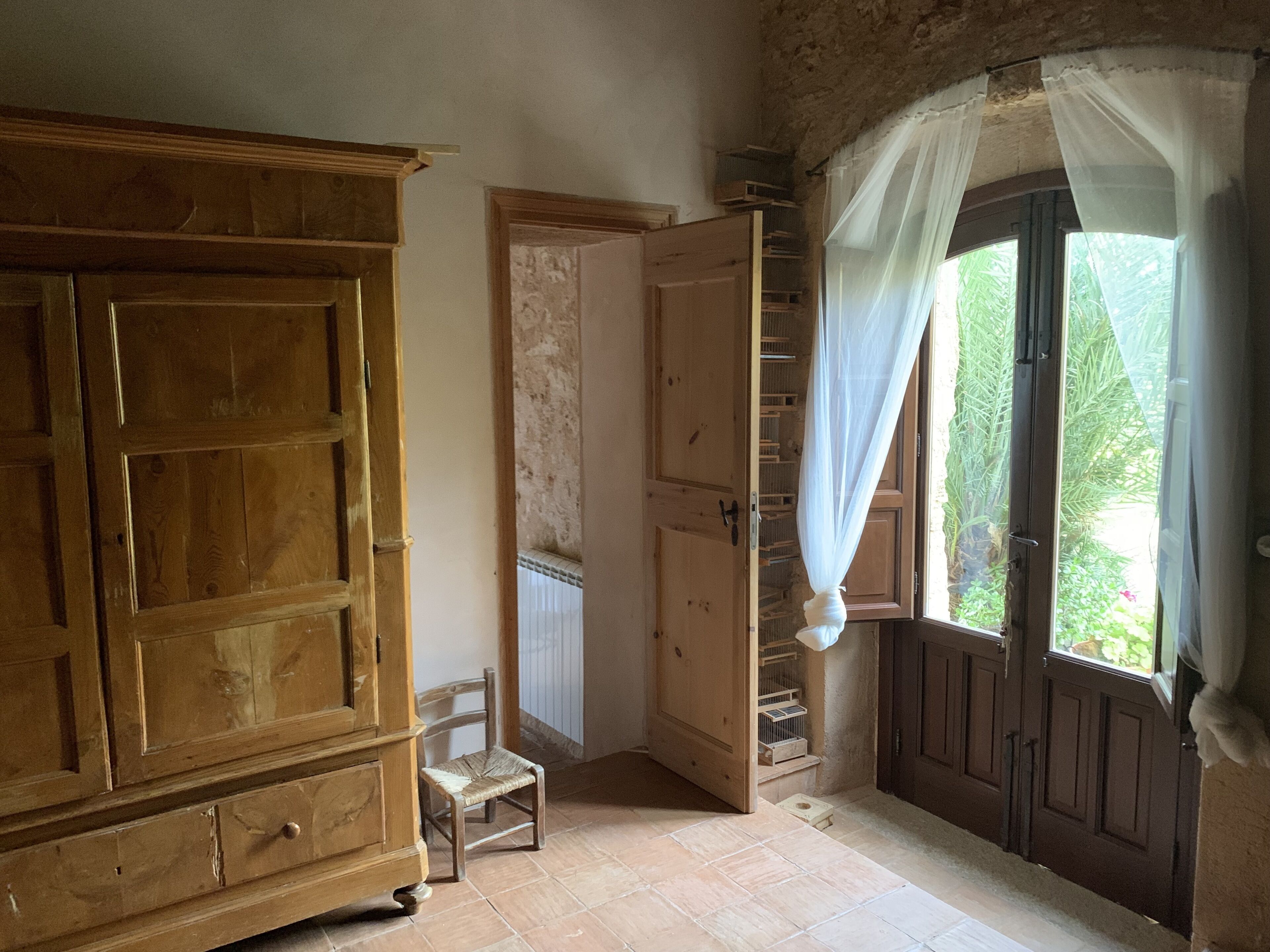 A room with a wooden cabinet, a small chair, a door leading to a balcony, and a window with curtains.