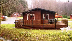 Una cabaña de madera con un deck cercado, rodeada de césped y un bosque.