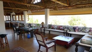 A spacious living area with a high ceiling, wooden furniture, a sofa set, a dining table, and a large window providing natural light.
