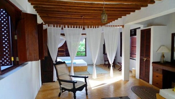 A room with a wooden ceiling, a bed, a chair, a desk, and a window with curtains.