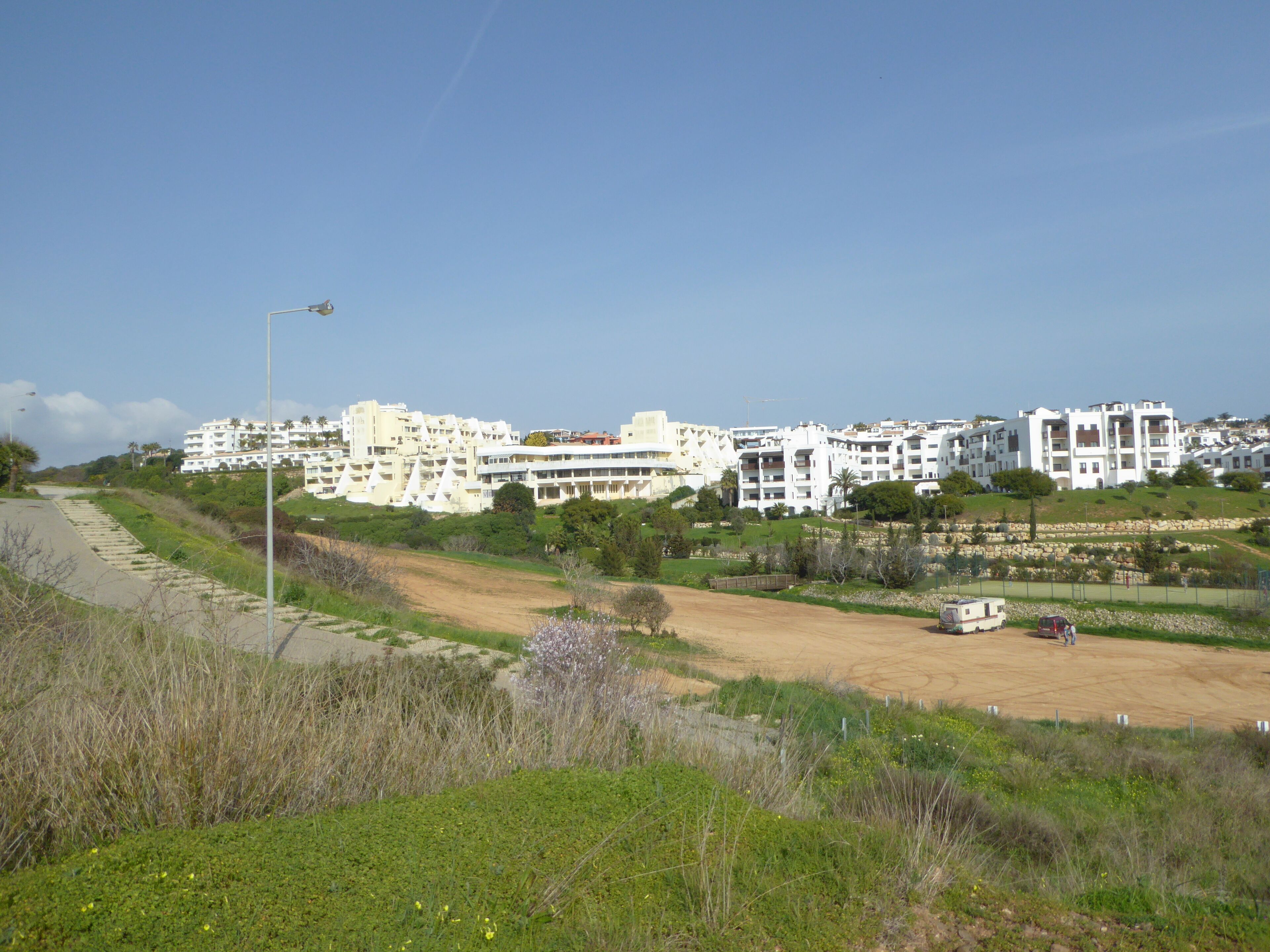 A coastal residential area with modern white buildings, a dirt path, and a parked camper.