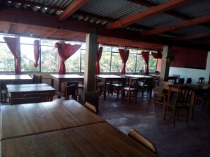 Un vaste espace dédié à la salle à manger, avec des tables et des chaises en bois, de grandes fenêtres ornées de rideaux rouges et un plafond haut avec des poutres apparentes.