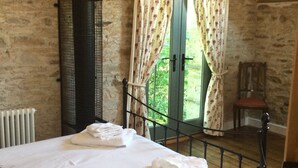 A bedroom with a stone wall, a bed with white linens, a radiator, a window with floral curtains, and a chair.