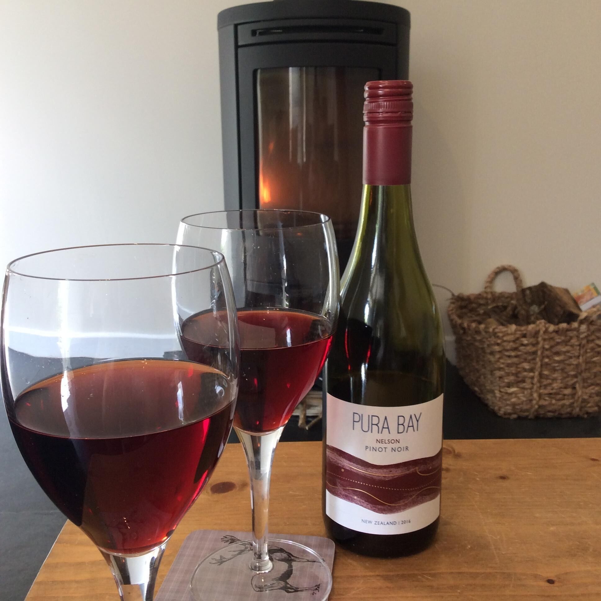 A bottle of Pura Bay Pinot Noir wine, two glasses of red wine, and a woven basket on a wooden table.