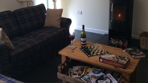 A cozy living room with a sofa, a wooden coffee table with chess and books, a fireplace, and a window with curtains.
