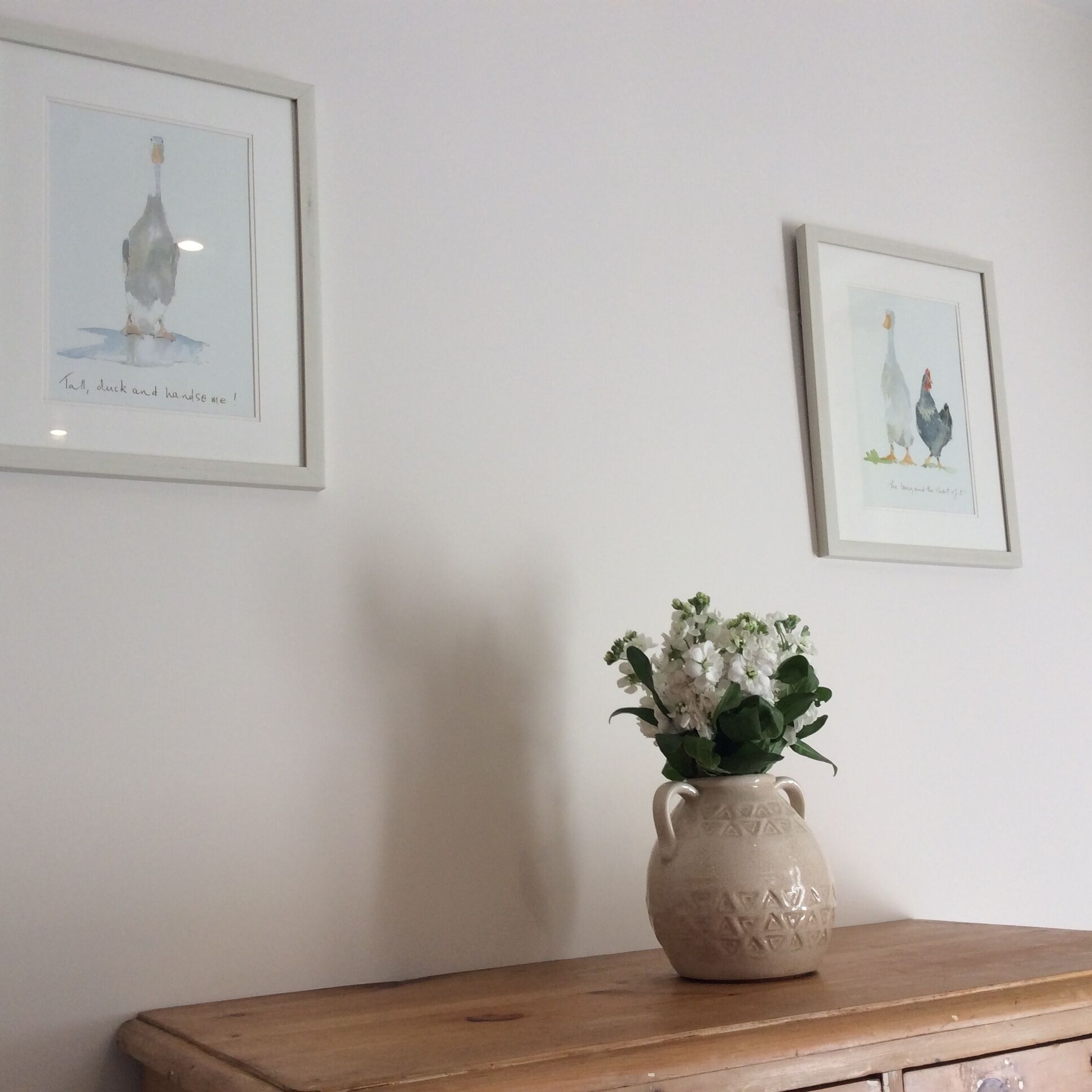 Une pièce comprenant une commode en bois, un vase avec des fleurs et deux œuvres d’art encadrées accrochées au mur.