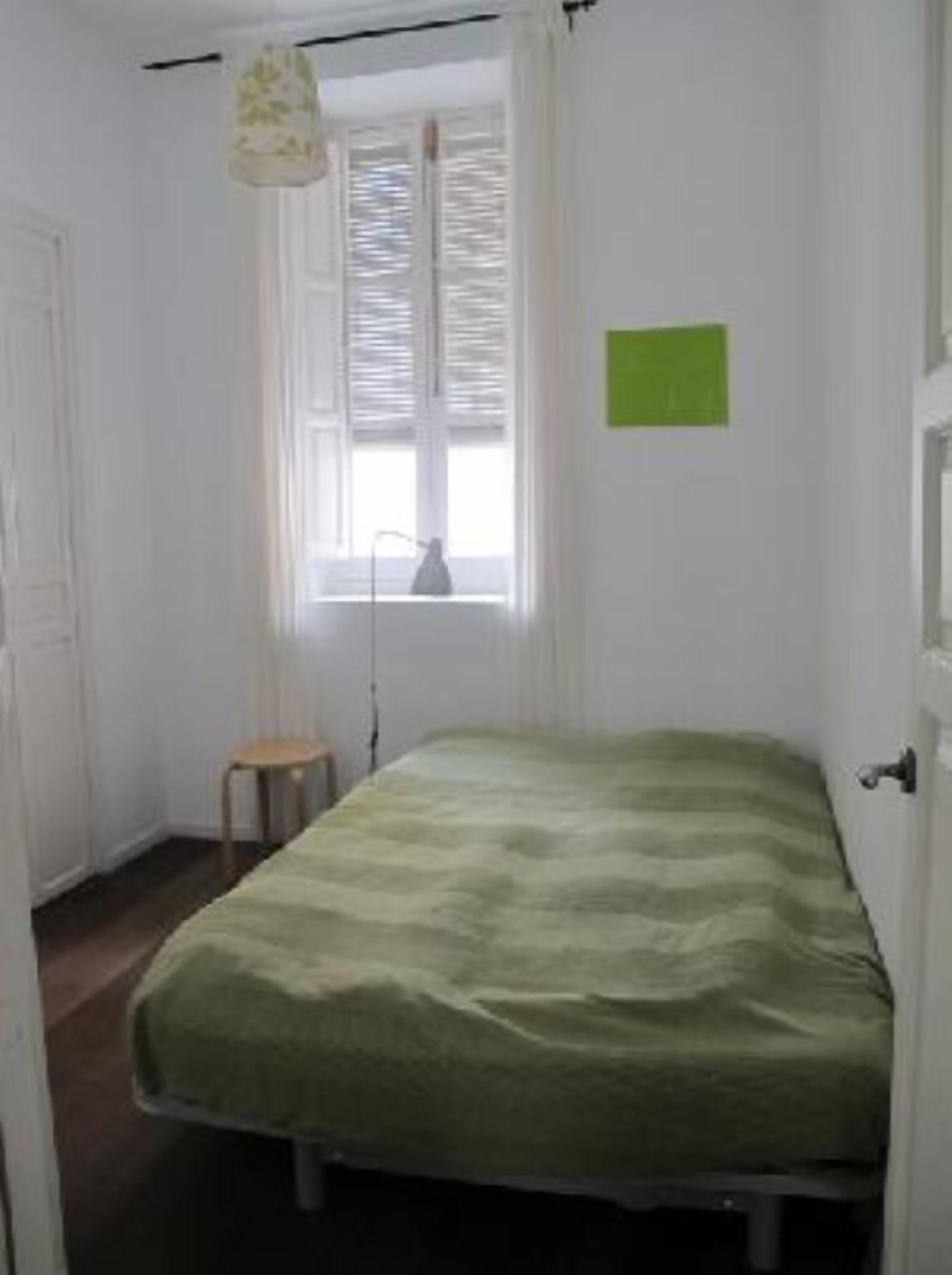 Una stanza piccola e pulita con un letto singolo, una finestra con persiane, un piccolo sgabello e un cartello verde appeso al muro.