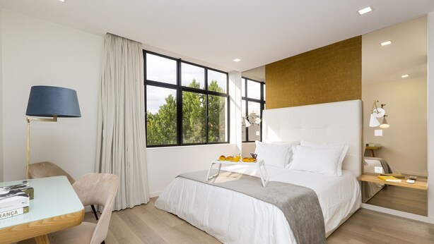 Ein modernes Hotelzimmer mit einem großen Bett, einem Schreibtisch mit Lampe, einem Sessel und Blick auf Bäume durch das Fenster.