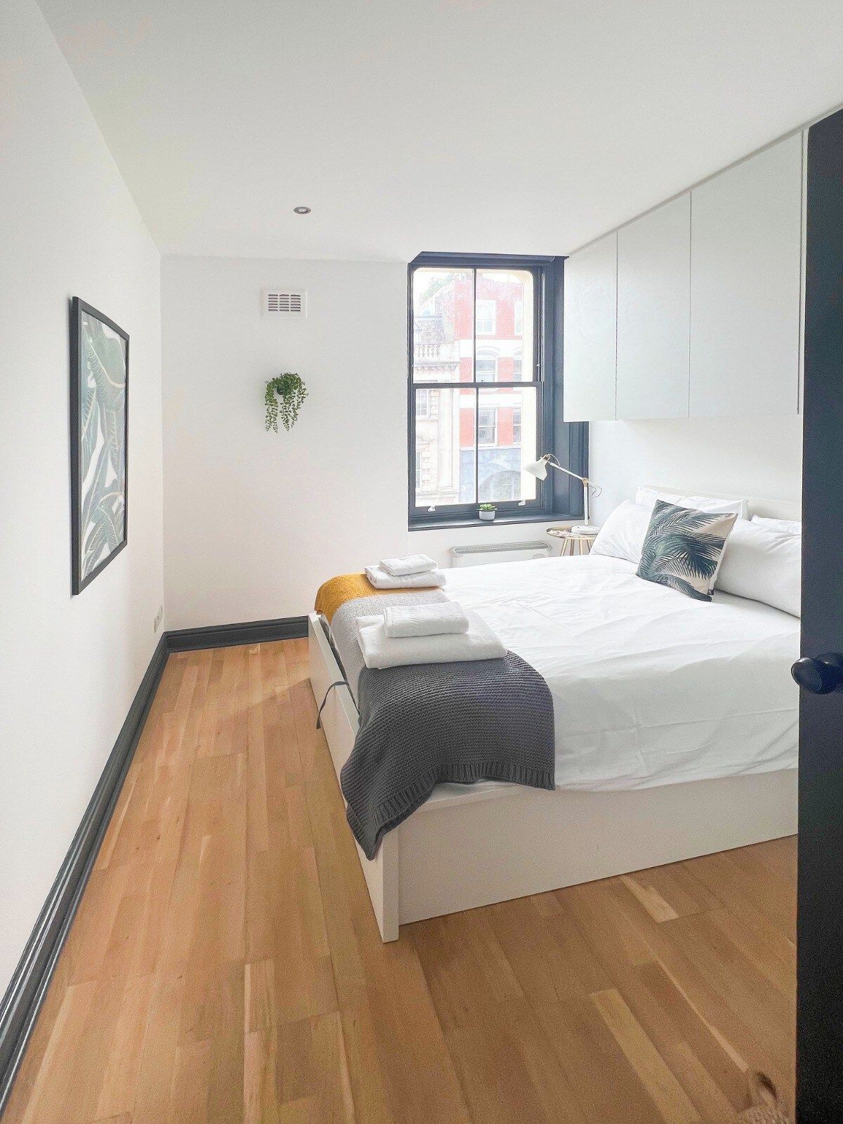 A modern bedroom with a large bed, a window with a view of buildings, a plant on the windowsill, and a framed picture on the wall.