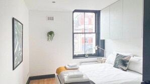 Un dormitorio moderno con una cama grande, un ventanal con vistas a edificios, una planta en el alféizar y un cuadro en la pared.