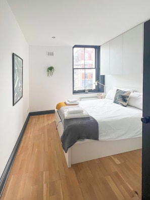 Un dormitorio moderno con una cama grande, un ventanal con vistas a edificios, una planta en el alféizar y un cuadro en la pared.