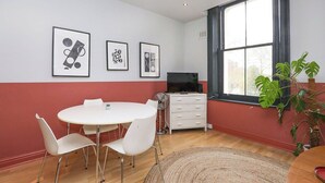 Una habitación con una mesa de comedor redonda, sillas blancas, un televisor de pantalla plana, un aparador blanco y un ventanal con vistas.
