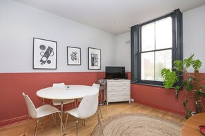 Una habitación con una mesa de comedor redonda, sillas blancas, un televisor de pantalla plana, un aparador blanco y un ventanal con vistas.