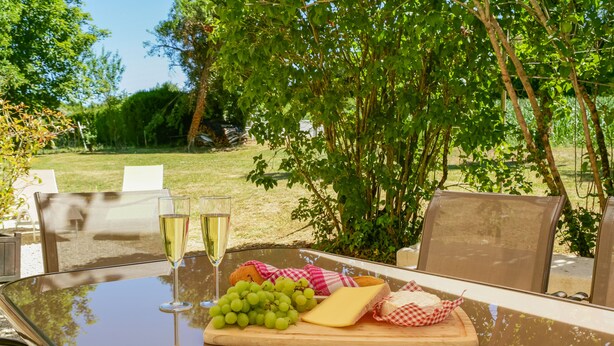 An outdoor dining table with a glass top, a wooden cutting board with grapes, cheese, and bread, and two glasses of white wine.