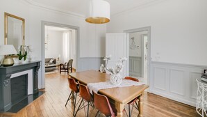 A dining area with a wooden table, chairs, and a white vase with flowers. There is a fireplace with a mirror and a lamp on the left.