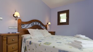 Quarto com cabeceira de madeira, cama com colcha florida, mesa de cabeceira com abajur e janela com vista para o verde.