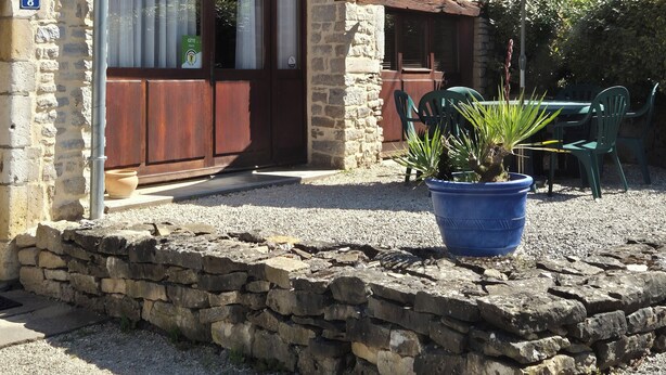 Il s’agit d’un bâtiment en pierre, doté d’un toit en tuiles, d’une porte en bois et d’une fenêtre avec des rideaux. Un parapluie bleu et un pot de fleurs se trouvent devant le bâtiment.