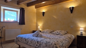 Une chambre à coucher avec un lit, deux lampes de chevet, une fenêtre avec des rideaux, un radiateur et une poutre en bois au plafond.