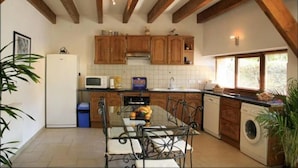 Une cuisine avec des meubles en bois, une table à manger avec des chaises, un lave-linge et une fenêtre avec des stores.