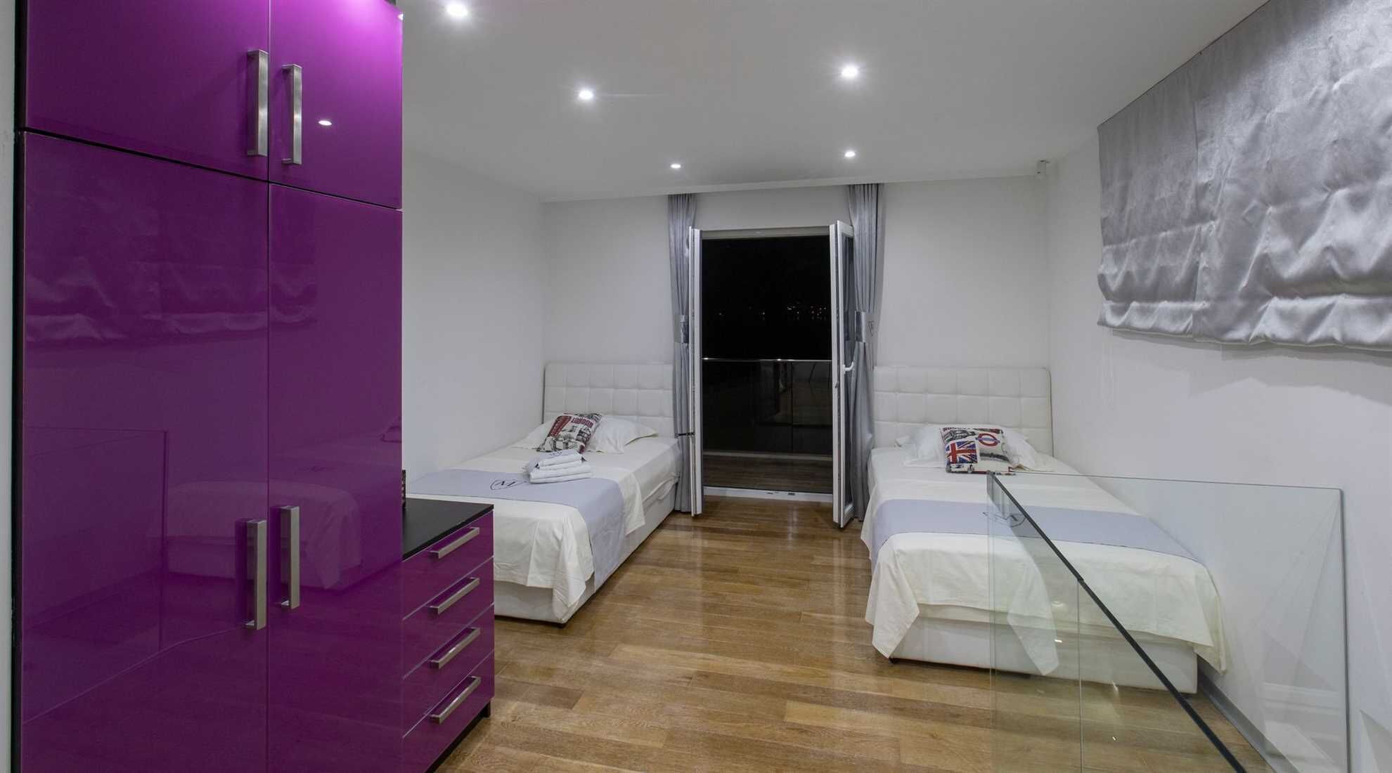 A modern bedroom with two beds, a purple wardrobe, and a large window.