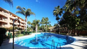 Piscina con una fuente de agua azul cristalina, rodeada de palmeras y un edificio con balcones.
