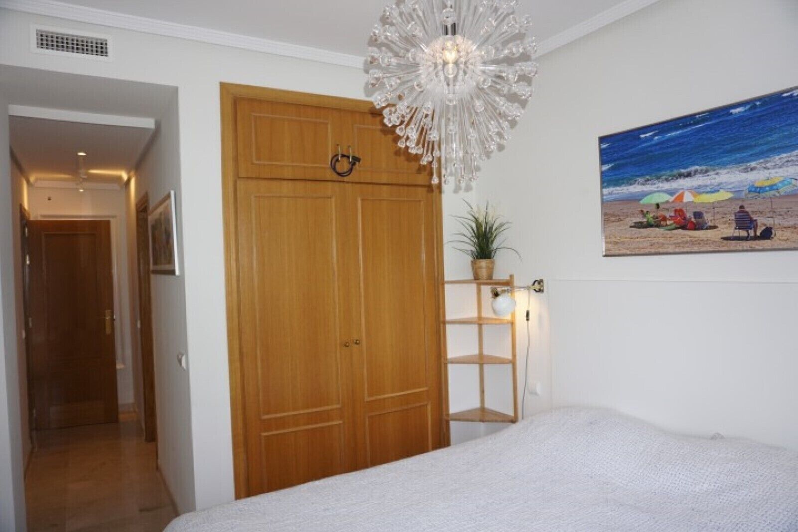 Ett sovrum med en garderob i trä, en vit säng, en hängande lampa, en hylla med en växt och en tavla med ett strandmotiv på väggen.