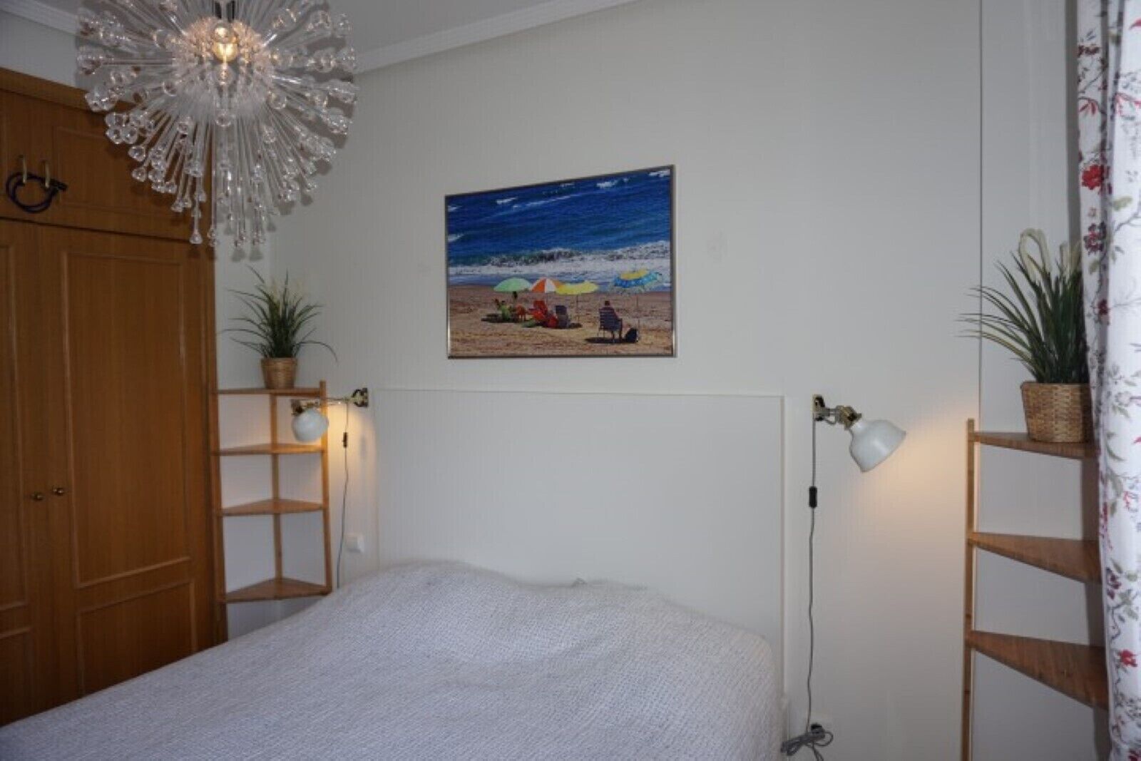 A bedroom with a bed, a wooden wardrobe, a chandelier, a painting of a beach scene, and two potted plants.