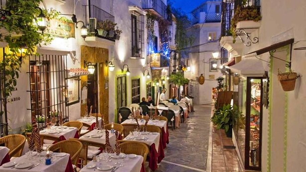 Un área de comedor al aire libre con mesas dispuestas para dos, rodeada de edificios con balcones y plantas colgantes.