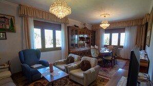 Een woonkamer met een blauwe fauteuil, twee beige fauteuils, een houten kast, een eettafel met stoelen en een televisie.