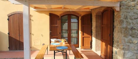 Un patio cubierto con mesa y bancos de madera, paredes de piedra y puertas de madera.