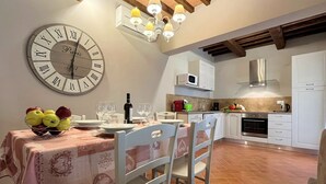 A dining area with a large wall clock, white chairs, a table set with plates, wine glasses, and a bowl of fruit.