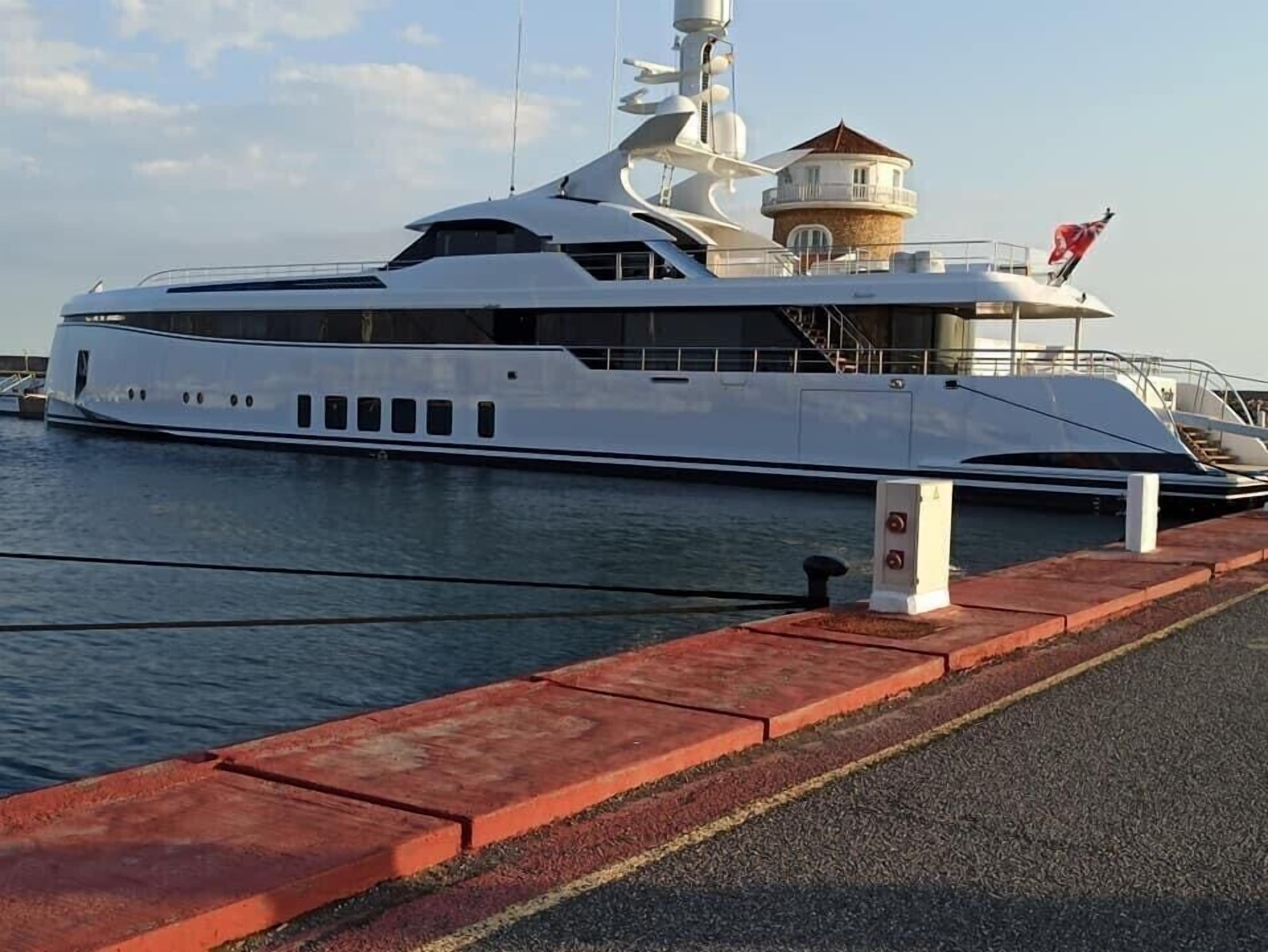A large white yacht docked at a marina with a red safety barrier and a small building structure on the dock.