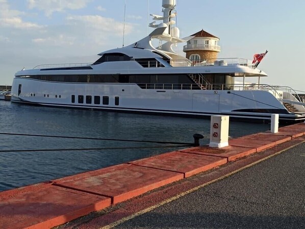 A large white yacht docked at a marina with a red safety barrier and a small building structure on the dock.