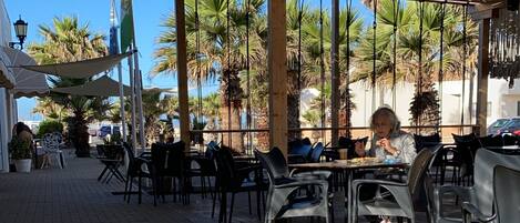 An outdoor café with brick flooring, metal chairs, and a shaded area with a metal roof.