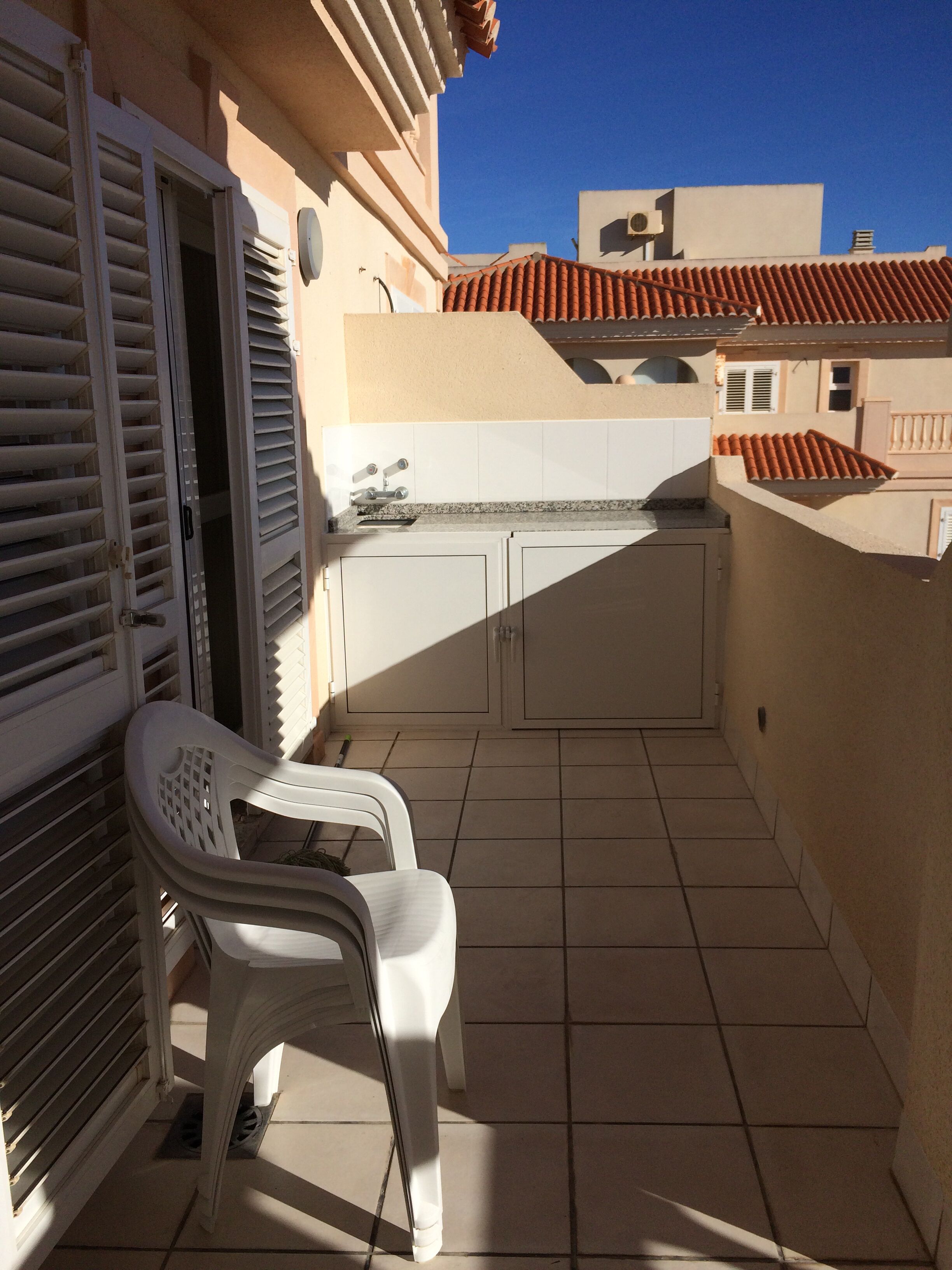A balcony with a white plastic chair, a small kitchen area with a sink and cabinets, and a tiled floor.