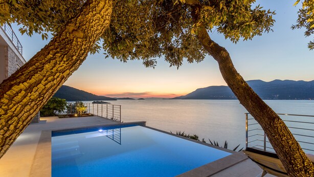 An infinity pool with a view of a calm lake and distant mountains at sunset. - Beachfront Villa Poseidon with Heated Pool (Korcula)