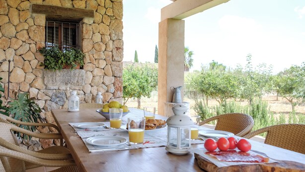 A stone wall with a window, a wooden table set for breakfast with plates, glasses, and a lantern, and a view of an orchard. - Nice relaxing country house with private pool (Inca)