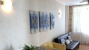 Ein modernes Wohnzimmer mit einem Glastisch, blauen Stühlen, einer Couch mit gemusterten Kissen und abstrakter Wandkunst.