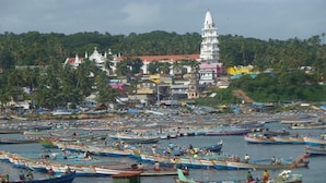 Une ville côtière dotée d’un grand port rempli de bateaux, d’une église blanche imposante surmontée d’une flèche, et d’une végétation luxuriante de palmiers en arrière-plan.