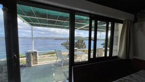 Habitación con un ventanal grande que da a una extensión de agua, un patio con mesa y sillas, y vistas al mar y la costa lejana.