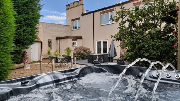 A hot tub in a backyard with a patio table and chairs, surrounded by greenery and a building in the background. - Large Family Cottage With Hot Tub, Ideally Located For Activities. Pet Friendly. (Newark)